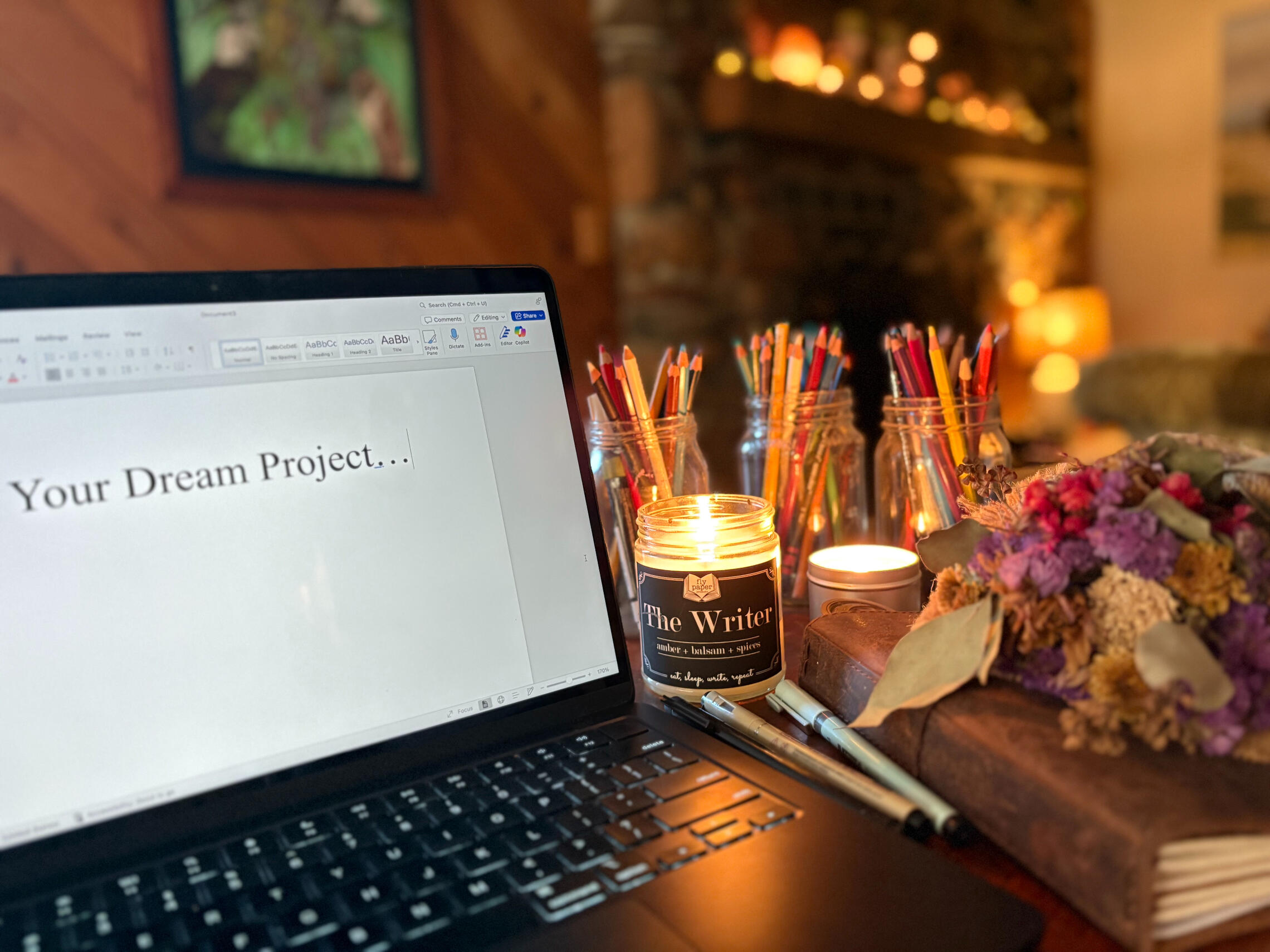 Photo: A computer surrounded by candles, a notebooks and pens, color pencils, and dried flowers. On the screen says: "Your Dream Project..."